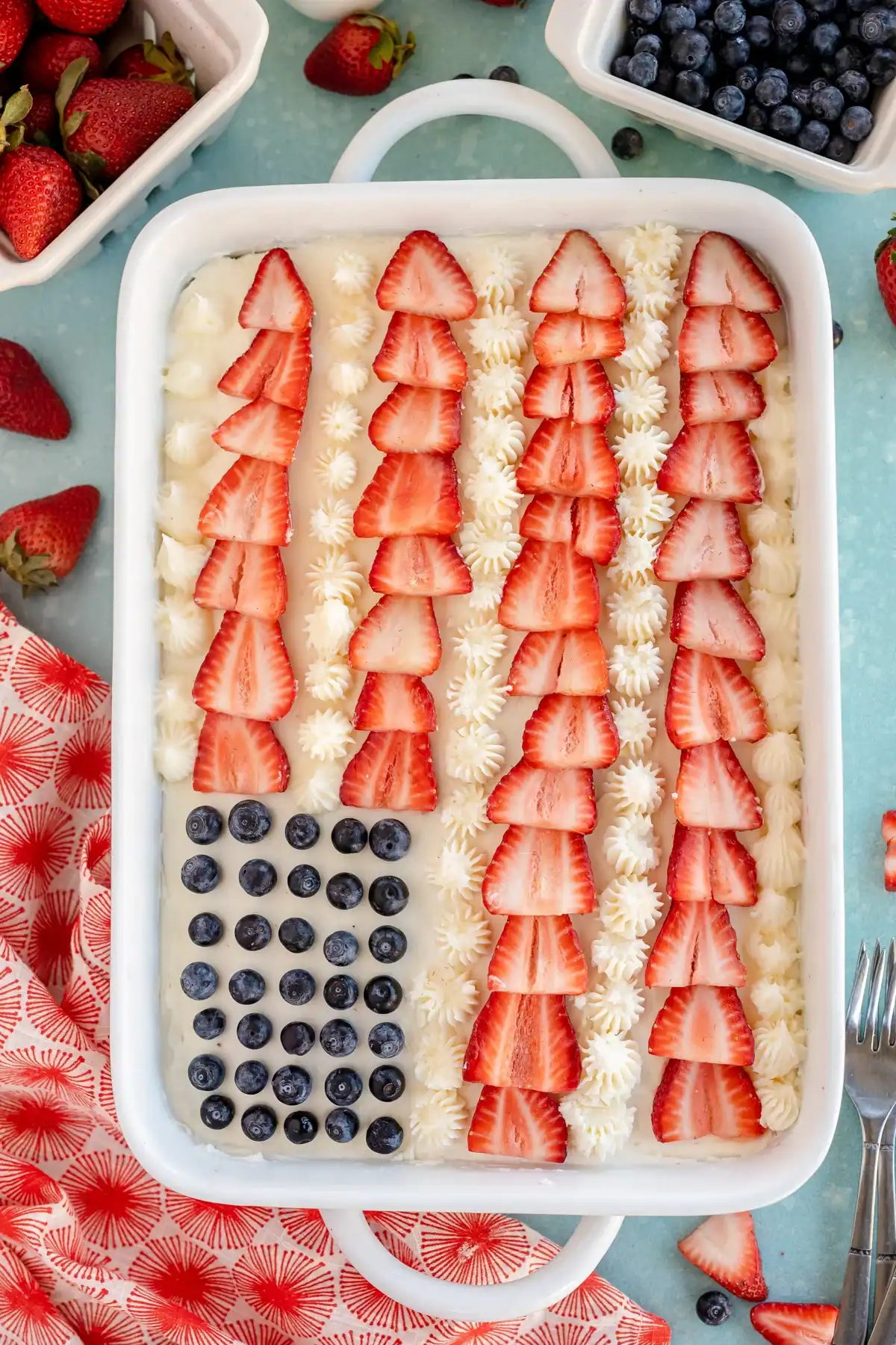 This festive dessert, inspired by classic 9x13 cake recipes, is decorated as an American flag with blueberries for stars, rows of sliced strawberries for stripes, and dollops of white cream in a white dish. Bowls of strawberries and blueberries are nearby, ready to add extra bursts of color and flavor.