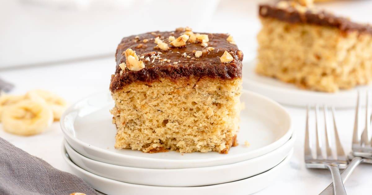 A slice of banana cake with chocolate frosting and chopped walnuts on top, served on a stack of white plates. Perfect for fans of 9x13 cake recipes, with another slice and sliced bananas in the background alongside a few forks.
