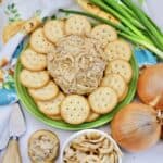 A plate with a French Onion Cheeseball coated in crispy fried onions, surrounded by round crackers. Nearby are whole onions, green onions, a bowl of fried onions, cheese spreaders, and a floral kitchen towel.