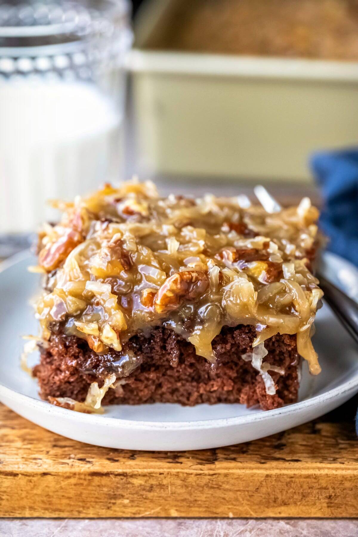 A close-up of a slice of chocolate cake from one of the best 9x13 cake recipes, topped with gooey coconut and pecan frosting, served on a white plate with a fork, plus a glass of milk and baking pan in the background.