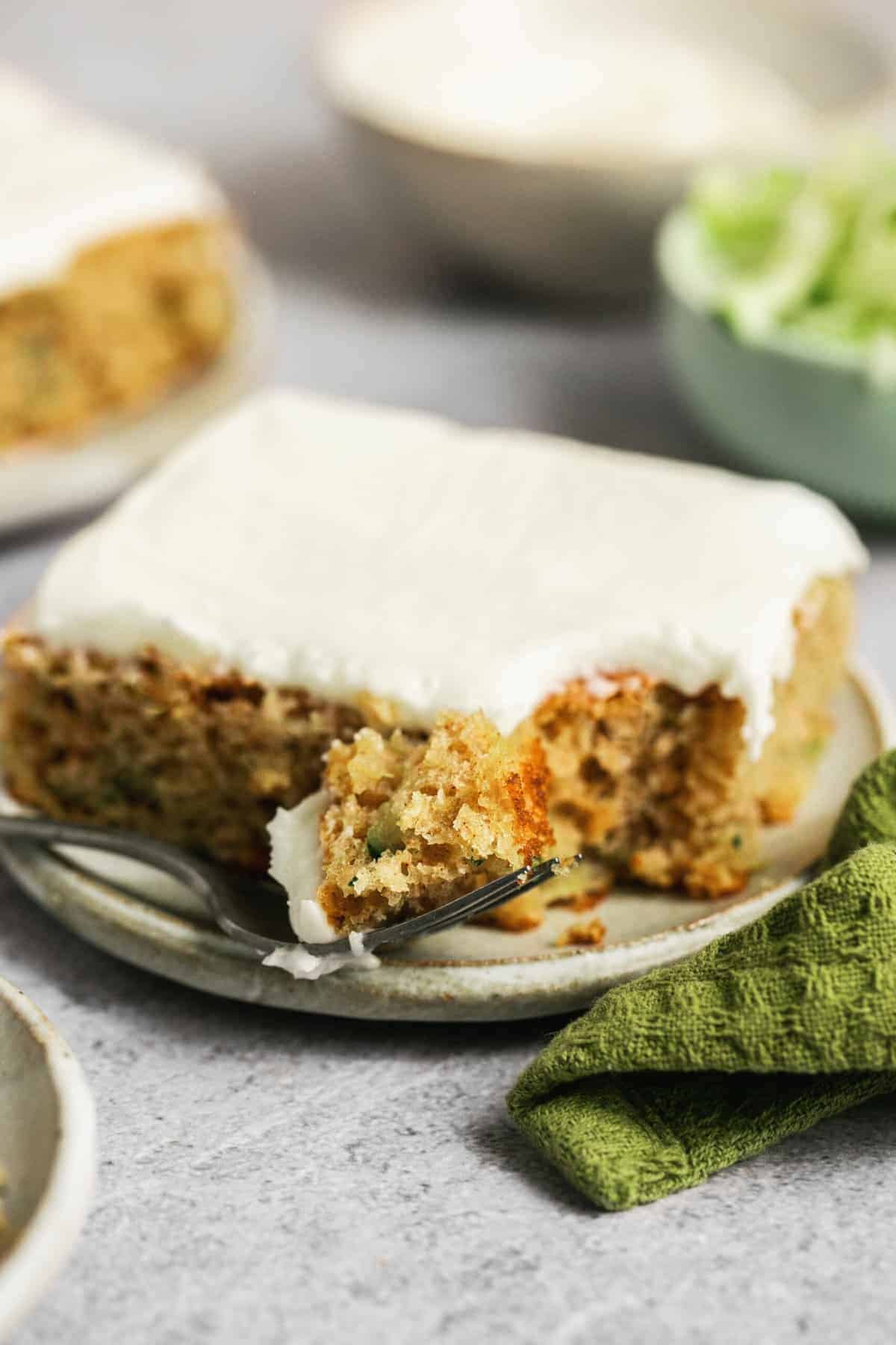 A slice of frosted cake from one of our favorite 9x13 cake recipes rests on a plate with a fork holding a bite. A green napkin lies beside the plate, and blurred dishes appear in the background.