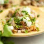 A close-up of a taco on a corn tortilla, filled with flavorful instant pot pork carnitas, chopped onions, and fresh cilantro, with another taco and extra cilantro visible in the background.