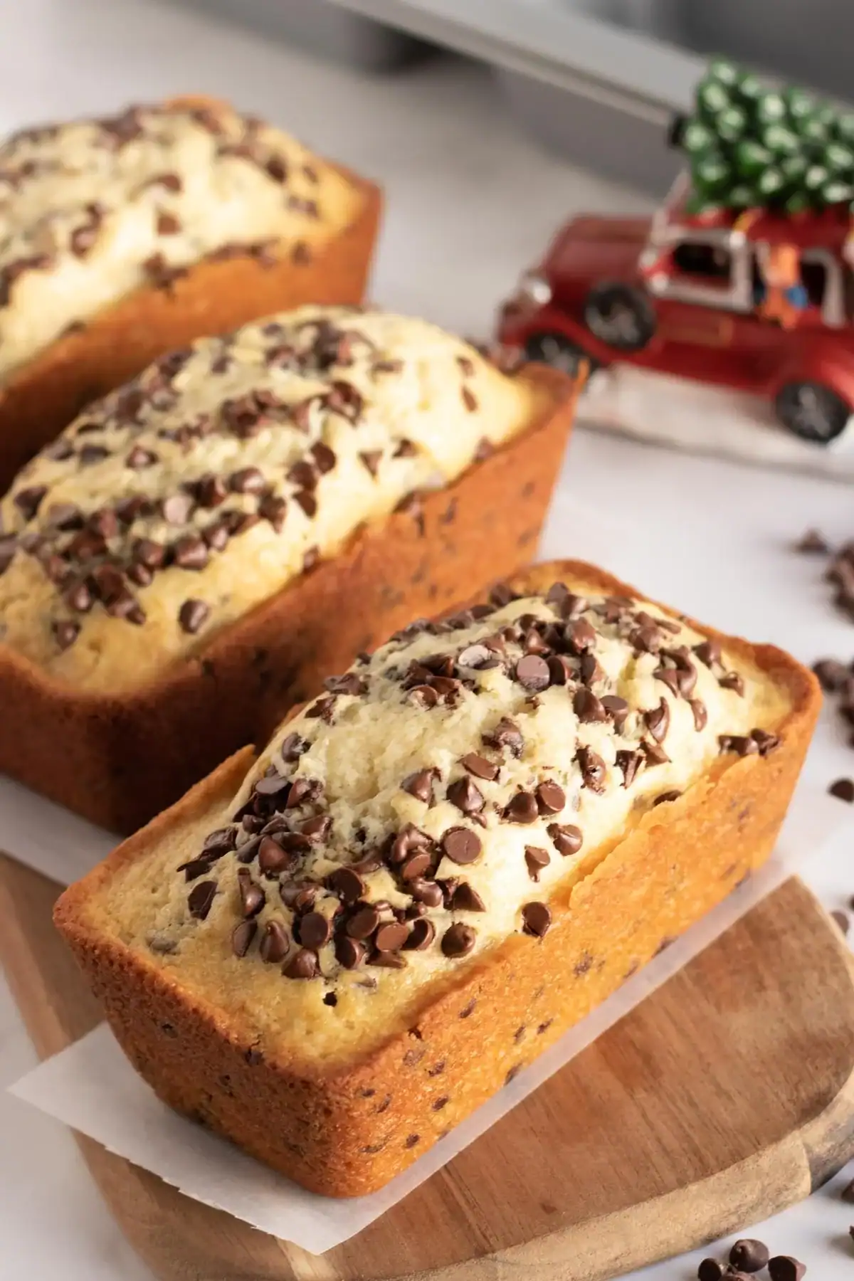Two mini loaves of chocolate chip bread rest on a wooden board, topped with chocolate chips—an irresistible answer to the question of what to make with chocolate chips besides cookies. In the background, a small red toy car with a Christmas tree on top adds a festive touch.