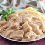 A white plate with creamy chicken and dumplings in gravy, served next to a portion of mashed potatoes and garnished with a sprig of parsley. A patterned cloth is partly visible in the background.