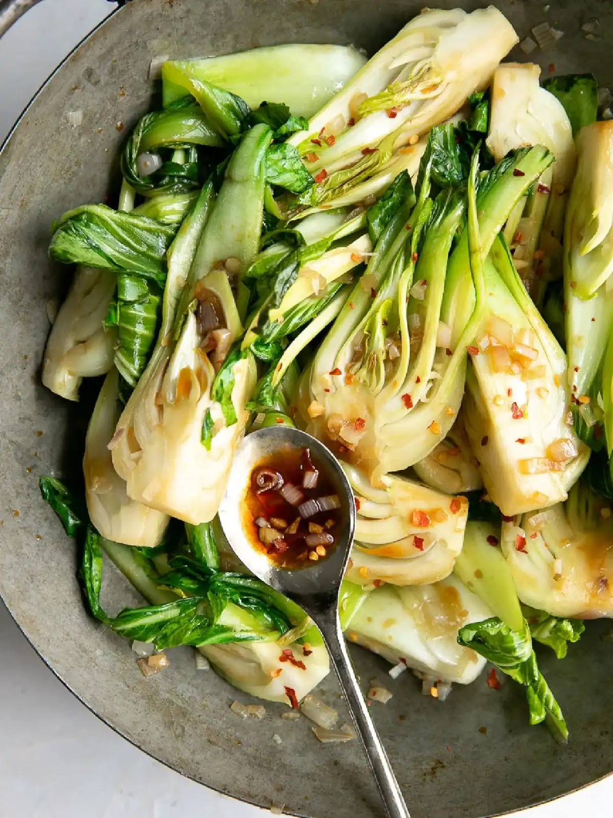 Sautéed bok choy cut in halves with red pepper flakes and a spoonful of soy-based sauce in a gray bowl, perfect for those seeking flavorful Thai side dishes.