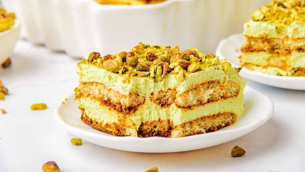 A slice of pistachio tiramisu, a standout among Green Desserts, sits on a white plate. The dessert features creamy layers and soaked ladyfinger biscuits, topped with chopped pistachios. A few nutty pieces are scattered on the table.