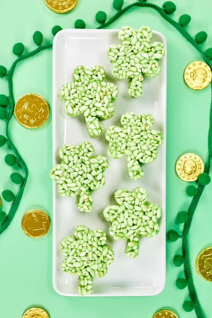A white rectangular plate holds six green clover-shaped crispy treats, perfect for anyone craving festive green desserts. The plate is surrounded by gold coins and a green pom-pom garland on a light green background.