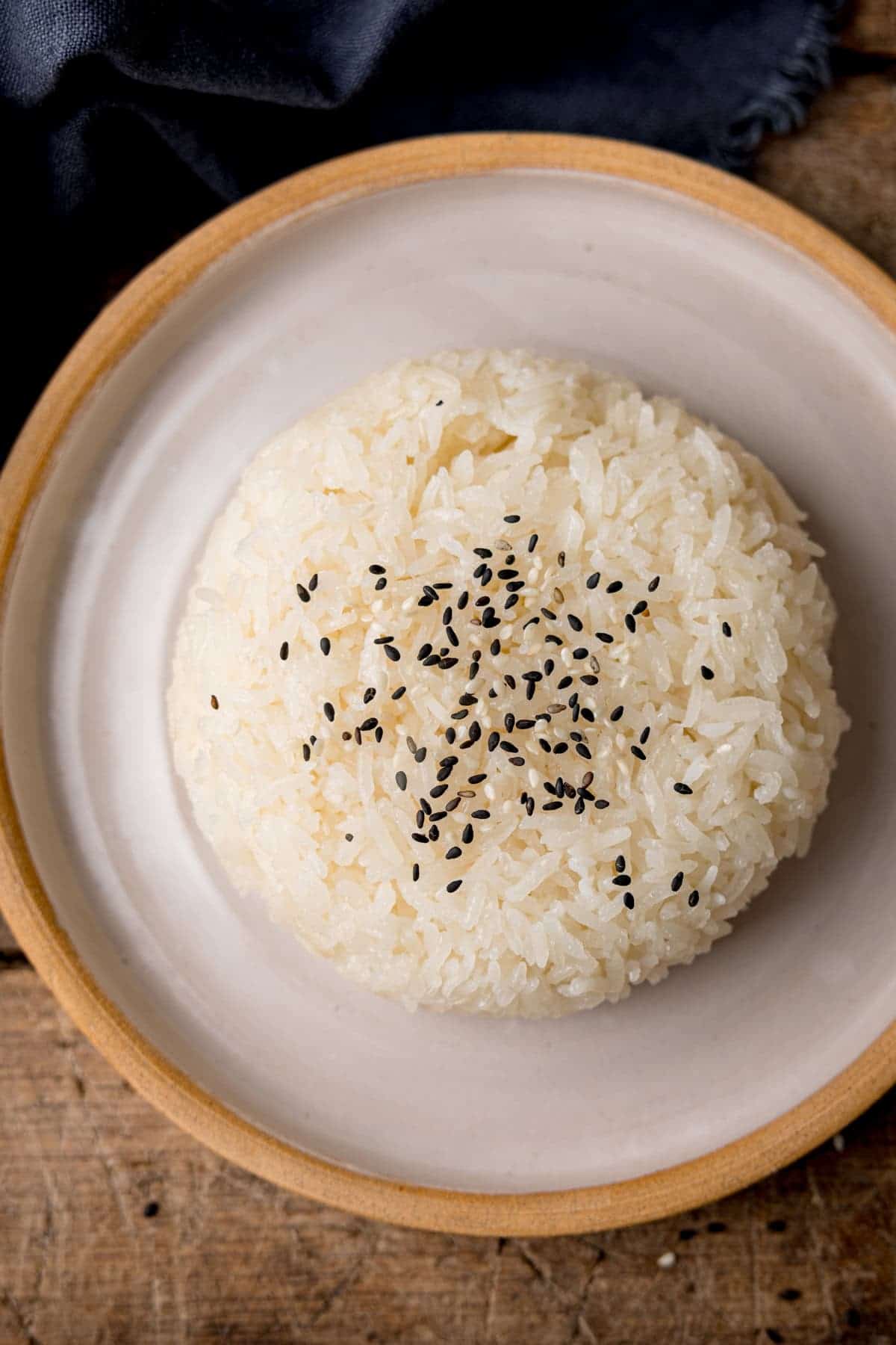 A round mound of white rice topped with black sesame seeds is served on a beige plate, set on a wooden surface with a dark cloth nearby—a simple yet classic presentation often found among Thai side dishes.