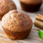 A close-up of a golden-brown Bisquick Muffin on a wooden surface, with cinnamon sticks and lime wedges nearby. More muffins are blurred in the background.