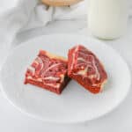 Two Red Velvet Cheesecake Brownies with cream cheese swirls are arranged on a white decorative plate, accompanied by a glass jar of milk and a soft white cloth in the background.