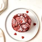A Red Velvet Crinkle Cookie dusted with powdered sugar sits on a white plate, surrounded by small red heart-shaped candies. A white cloth is partially visible on the side, adding charm to this sweet Red Velvet Crinkle Cookies display.