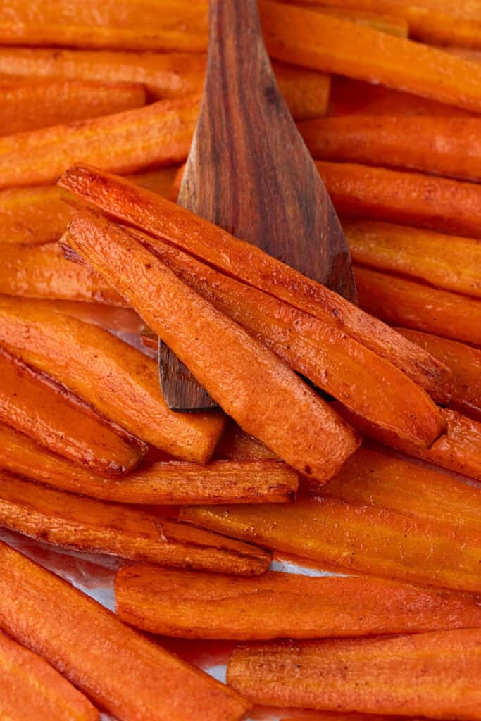 A close-up of Baked Carrots with Brown Sugar on a tray, with a wooden spatula lifting a few pieces. The carrots are golden-brown and glossy, indicating they are well-cooked.