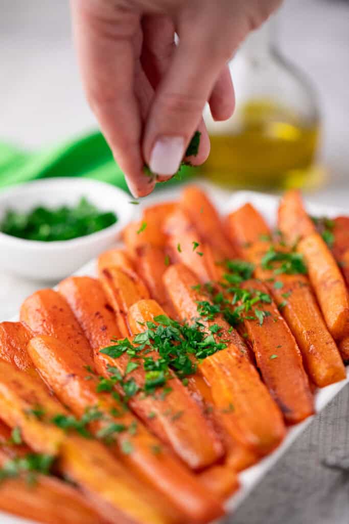 A hand sprinkles chopped fresh herbs over a platter of Baked Carrots with Brown Sugar, with a bowl of herbs and a bottle of oil visible in the background.