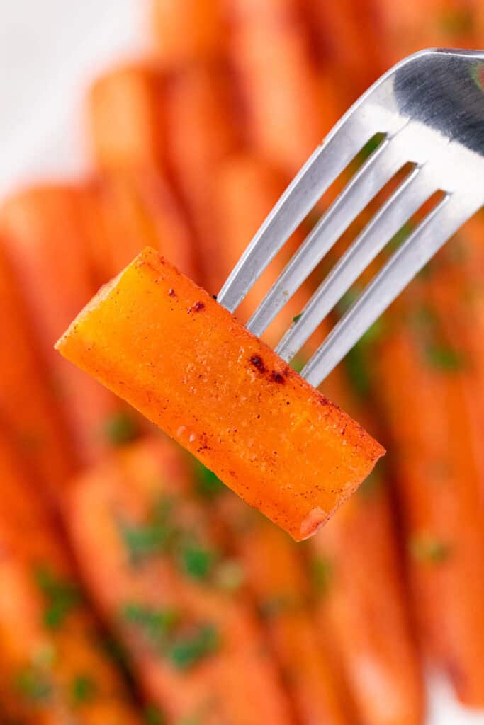 A close-up of a fork holding a seasoned, roasted carrot stick, with more baked carrots with brown sugar blurred in the background. The carrot appears to be sprinkled with herbs and spices.