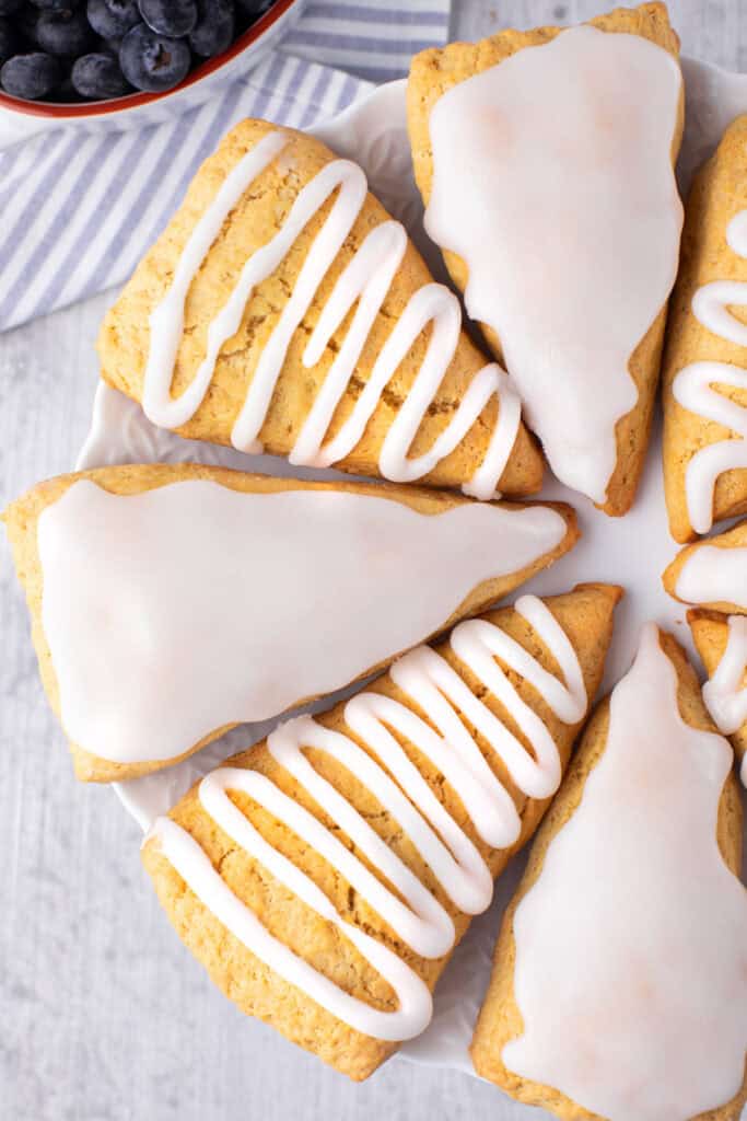 A plate of golden brown Bisquick scones topped with white icing, arranged in a circle on a white plate. A bowl of blueberries is partially visible in the upper left corner. The background is a light wooden surface.