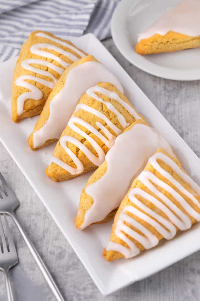 Four iced Bisquick scones with white glaze are arranged on a rectangular white plate. A fork is nearby, and a plate with another glazed scone is partially visible in the background. The scene is set on a light gray surface.