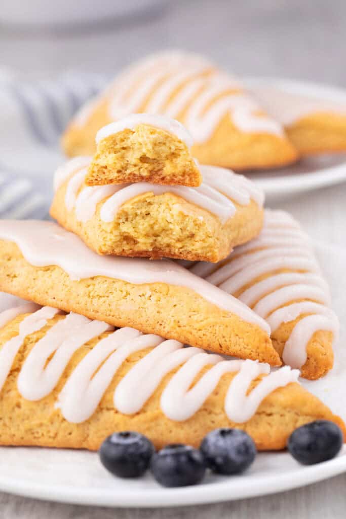 Three triangular bisquick scones with white icing are stacked on a plate, one broken in half to reveal its fluffy texture. Blueberries are scattered around, and more scones can be seen in the background.