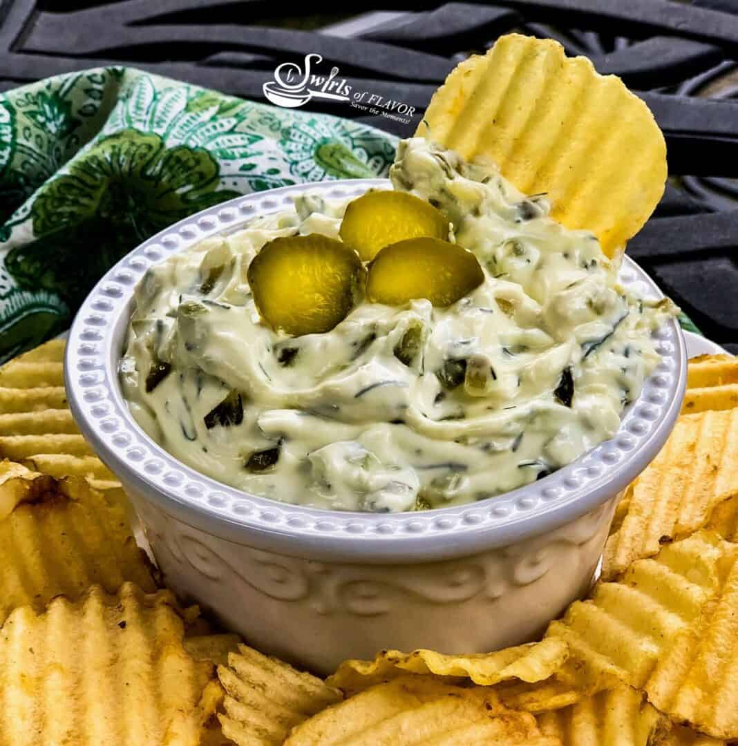 A white bowl filled with creamy dip topped with sliced pickles, surrounded by ridged potato chips, sits on a textured black surface with a green patterned cloth in the background.