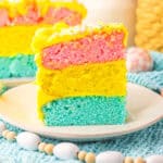 A slice of colorful Easter layer cake on a white plate. The cake has three vibrant layers—blue on the bottom, yellow in the middle, and pink on top—frosted with bright yellow icing and topped with pastel sprinkles. The background features spring-themed decorations including a blue crocheted placemat, pastel gingham eggs, wooden beads, and a white ceramic vase with greenery.