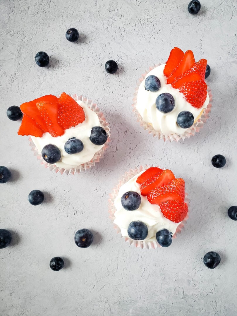 Three cupcakes with white frosting, topped with fresh strawberry slices and blueberries, are arranged on a light gray textured surface with scattered blueberries—perfect for Memorial Day dessert recipes.