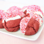 A plate of red velvet cookies dipped in white chocolate and pink sprinkles.