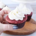 A hand with glittery blue nail polish holds a red velvet cupcake topped with white swirled frosting. More red velvet cupcakes with frosted tops are arranged on a wooden board in the background.
