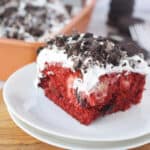 A slice of Red Velvet Poke Cake with Oreos, topped with white whipped topping and crumbled chocolate cookies, sits on a white plate, while the rest of the cake waits in a baking dish in the background.