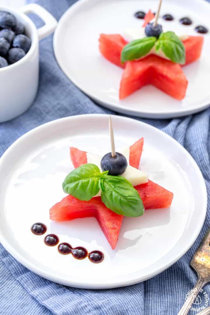 A white plate with a star-shaped watermelon slice, topped with a blueberry and basil leaves, secured by a toothpick. Balsamic glaze dots are on the side. Another similar plate and a cup of blueberries are in the background.