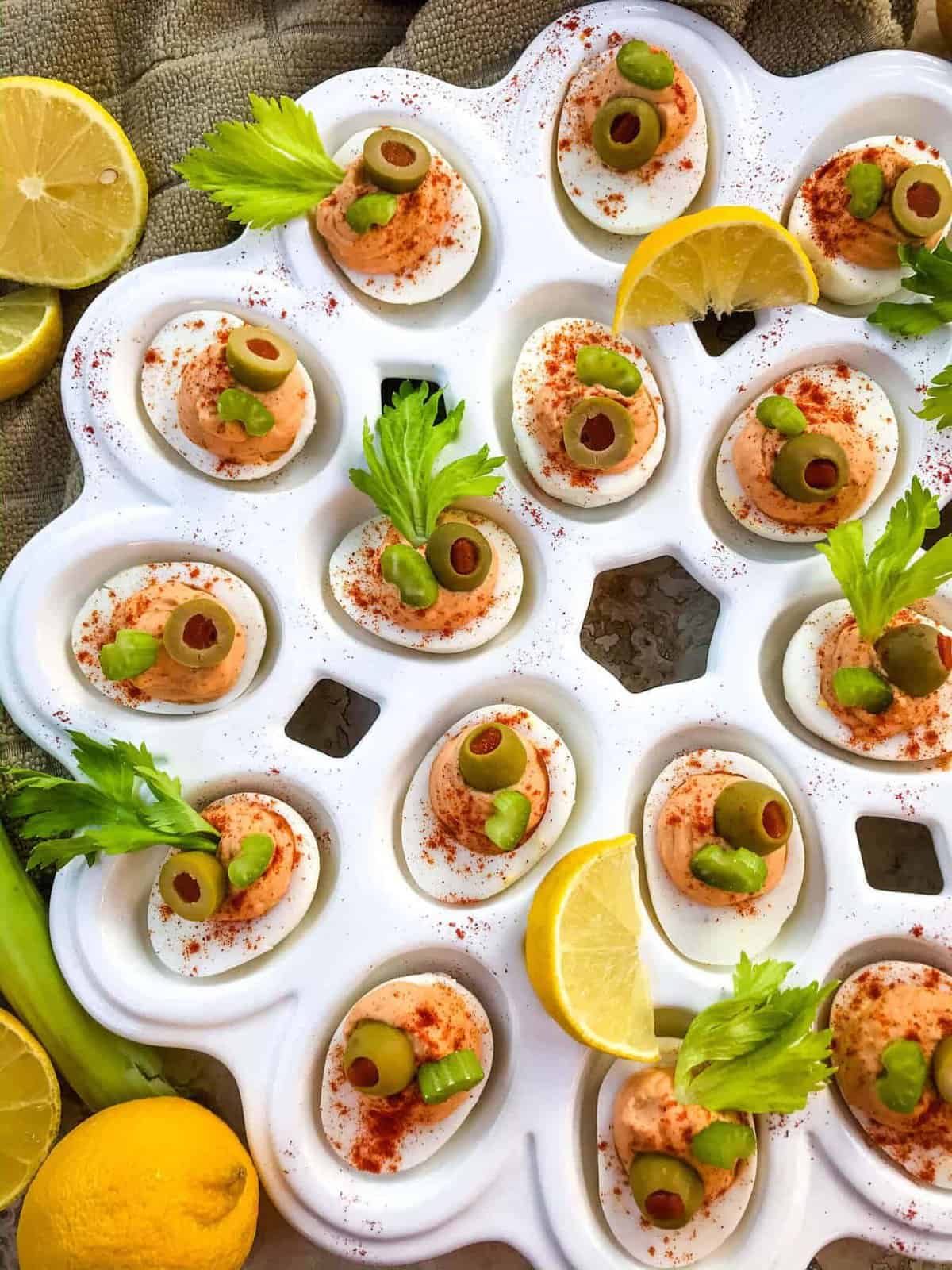 A tray of deviled eggs topped with green olives, celery leaves, and paprika makes perfect Spring Finger Foods. Lemon wedges and fresh celery garnish the neatly arranged eggs on a white serving dish.