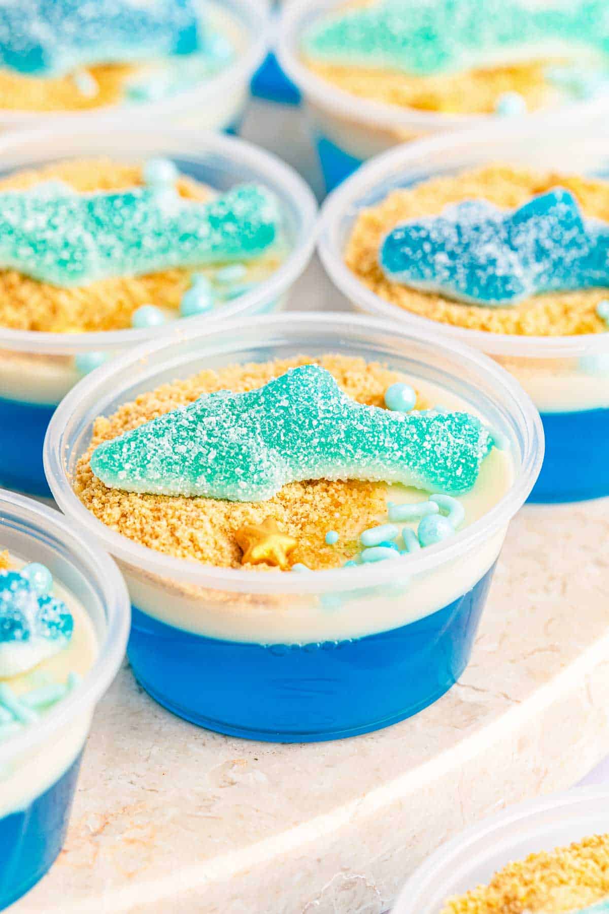 Clear plastic cup filled with blue gelatin, topped with crushed cookie crumbs, white cream, blue and white candy pearls, and a blue gummy candy shaped like a shark—an eye-catching shark snack. Similar cups are visible in the background.