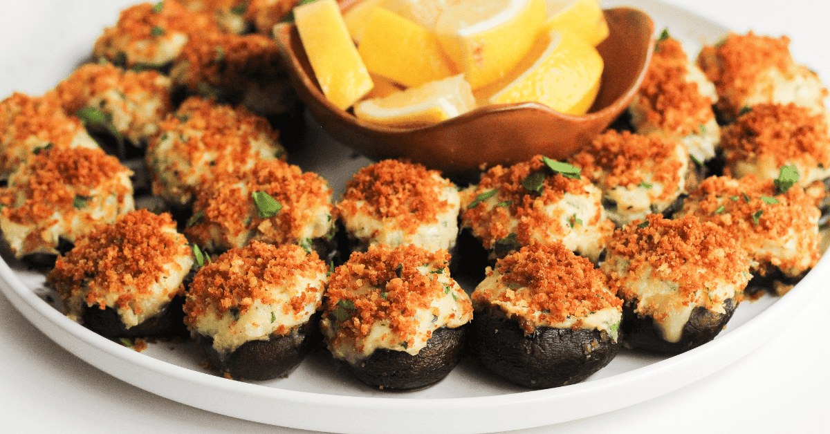 A plate of stuffed mushrooms topped with golden breadcrumbs is arranged around a small bowl filled with lemon wedges. Garnished with chopped herbs, these Spring Finger Foods are perfect for any seasonal gathering.