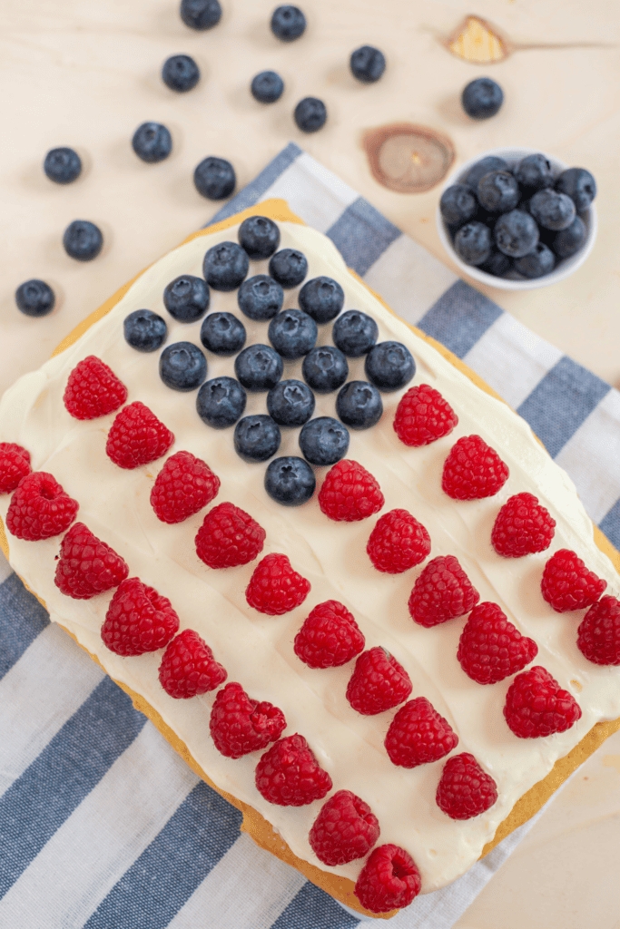 A rectangular cake with white frosting is decorated using Red White & Blue Cake Decorating Ideas—rows of raspberries and a corner of blueberries mimic the American flag. The cake rests on a blue and white striped cloth, with extra blueberries nearby.