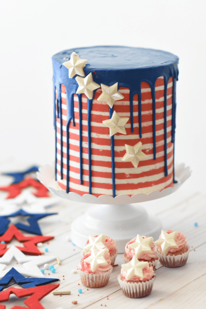 A tall cake with red and white stripes, blue drip icing, and white chocolate stars sits on a cake stand. Three cupcakes with pink frosting and white stars are in front—perfect for Red White & Blue Cake Decorating Ideas. Star decorations complete the scene.