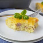A slice of cheesy breakfast casserole recipe with ham on a white plate, garnished with fresh basil leaves, with a fork beside it and another plate in the background.