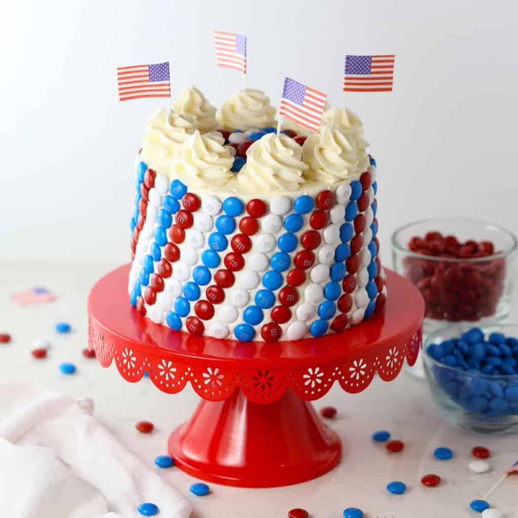 A cake decorated with red, white, and blue M&M candies and small American flags sits on a red cake stand—perfect for anyone seeking Red White & Blue Cake Decorating Ideas. Bowls of colorful candies complete the festive scene.