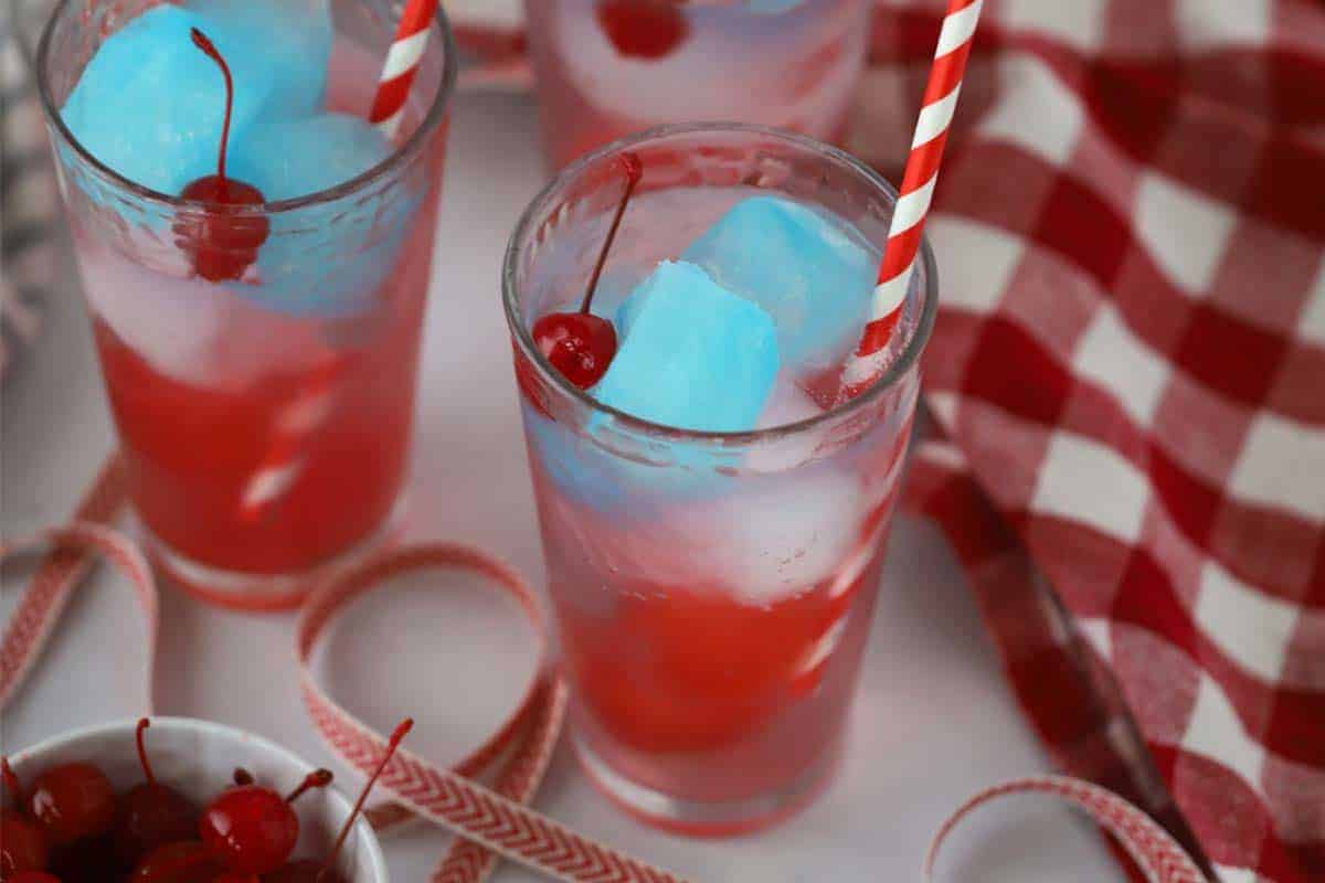 Three glasses of Fourth of July non alcoholic drinks with blue ice cubes, garnished with cherries and red striped straws. A bowl of cherries and a red-and-white checkered cloth are nearby.