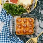 A glass baking dish with nine meatball sliders—slider rolls topped with meatballs, marinara sauce, and cheese—sits on a marble surface next to fresh parsley, grated cheese, a gold spatula, and a blue checkered cloth.
