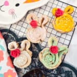 Four Easter Bunny Cinnamon Rolls with pastel icing and red rose decorations sit on a cooling rack. A floral napkin and a bunny-themed plate are nearby, creating a festive and colorful Easter scene.