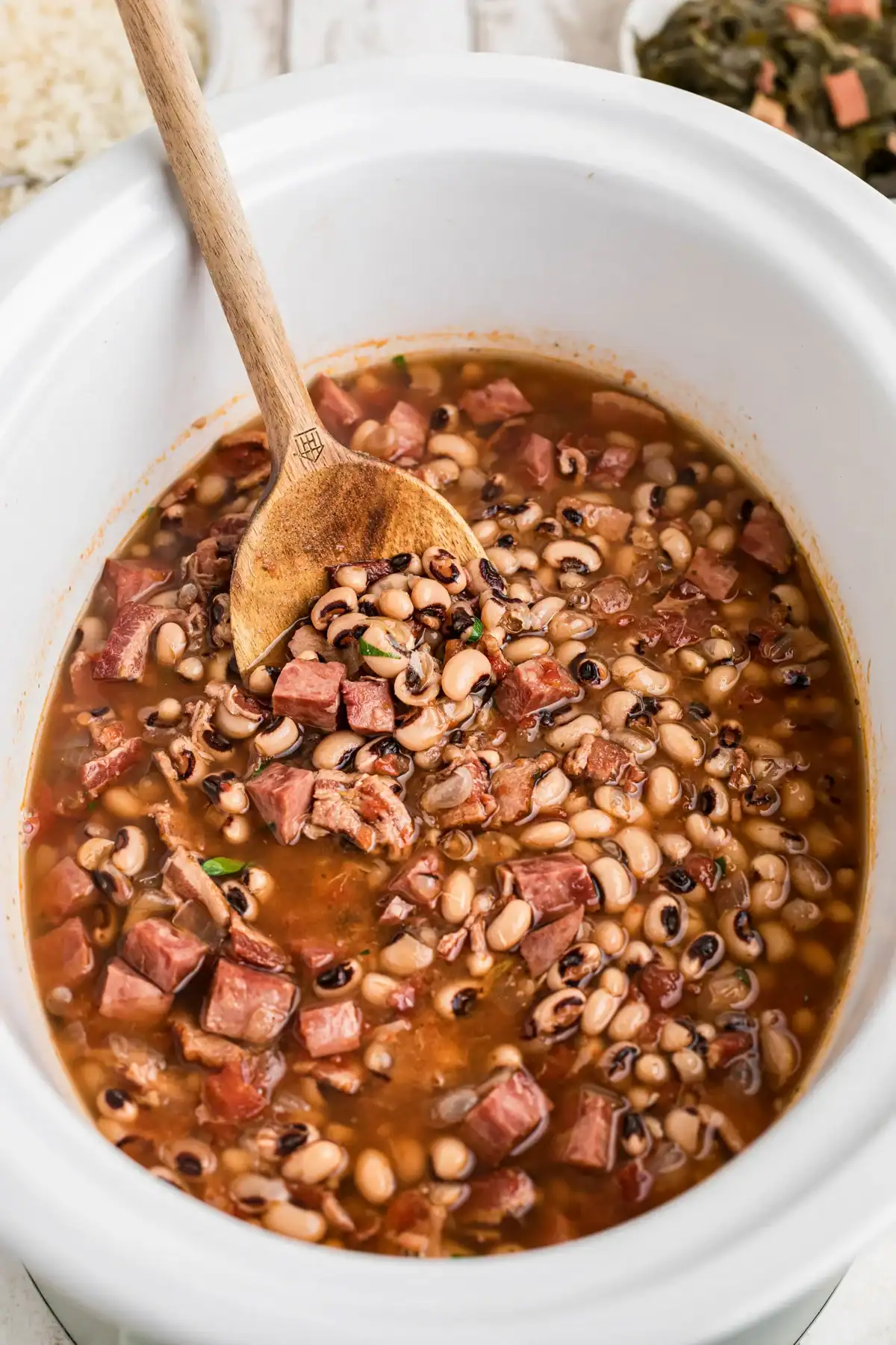 A wooden spoon stirs a hearty mixture of black-eyed peas, chopped ham, and broth in a white slow cooker—one of the perfect Slow Cooker Side Dishes for BBQ gatherings.