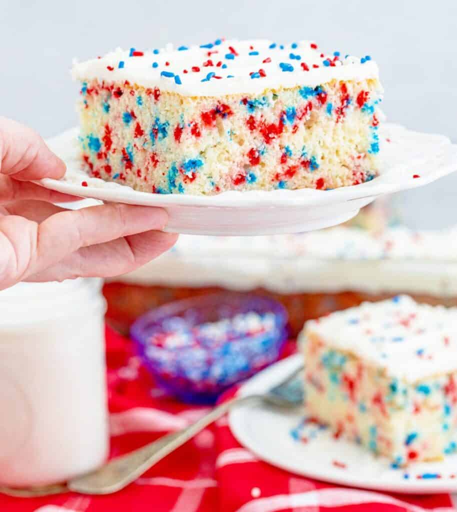 A hand holds a plate with a slice of white cake topped with white frosting and red, white, and blue sprinkles—perfect for anyone seeking Red White & Blue Cake Decorating Ideas. The cake's vibrant red and blue spots add to its festive appeal.