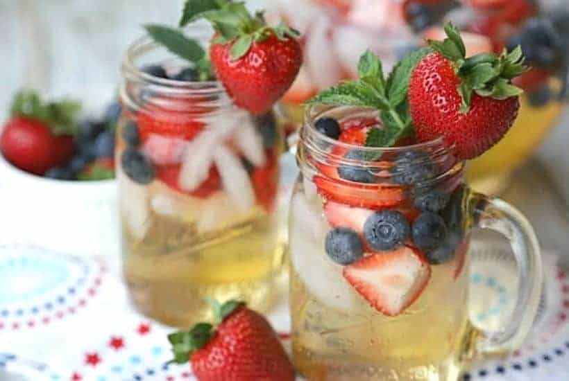 Two mason jars filled with a light-colored Fourth of July non alcoholic drink, ice, sliced strawberries, blueberries, and garnished with whole strawberries and mint leaves sit on a festive tablecloth with red and blue stars.