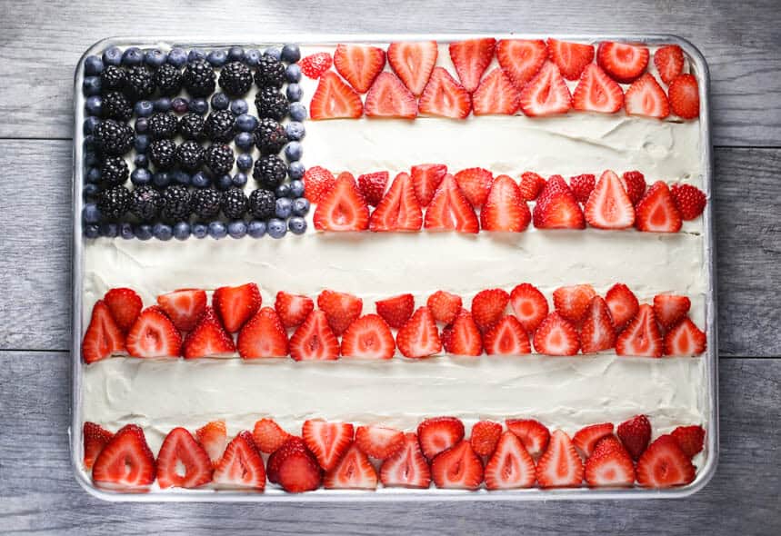 A sheet cake decorated as an American flag with blueberries and blackberries for the stars area and rows of sliced strawberries for the stripes on white frosting—perfect inspiration for Red White & Blue Cake Decorating Ideas, displayed on a wooden surface.