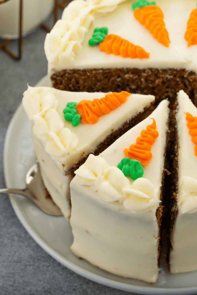 A round carrot cake with cream cheese frosting, decorated with small orange and green icing carrots on top—a charming example of carrot cake decorating ideas. One slice is being lifted from the cake with a cake server.