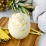 A glass of creamy pineapple dessert sits on a wooden board, garnished with two yellow-striped straws and pineapple slices, evoking the classic Disney Dole Whip Recipe. A whole pineapple and a white cloth are in the background.
