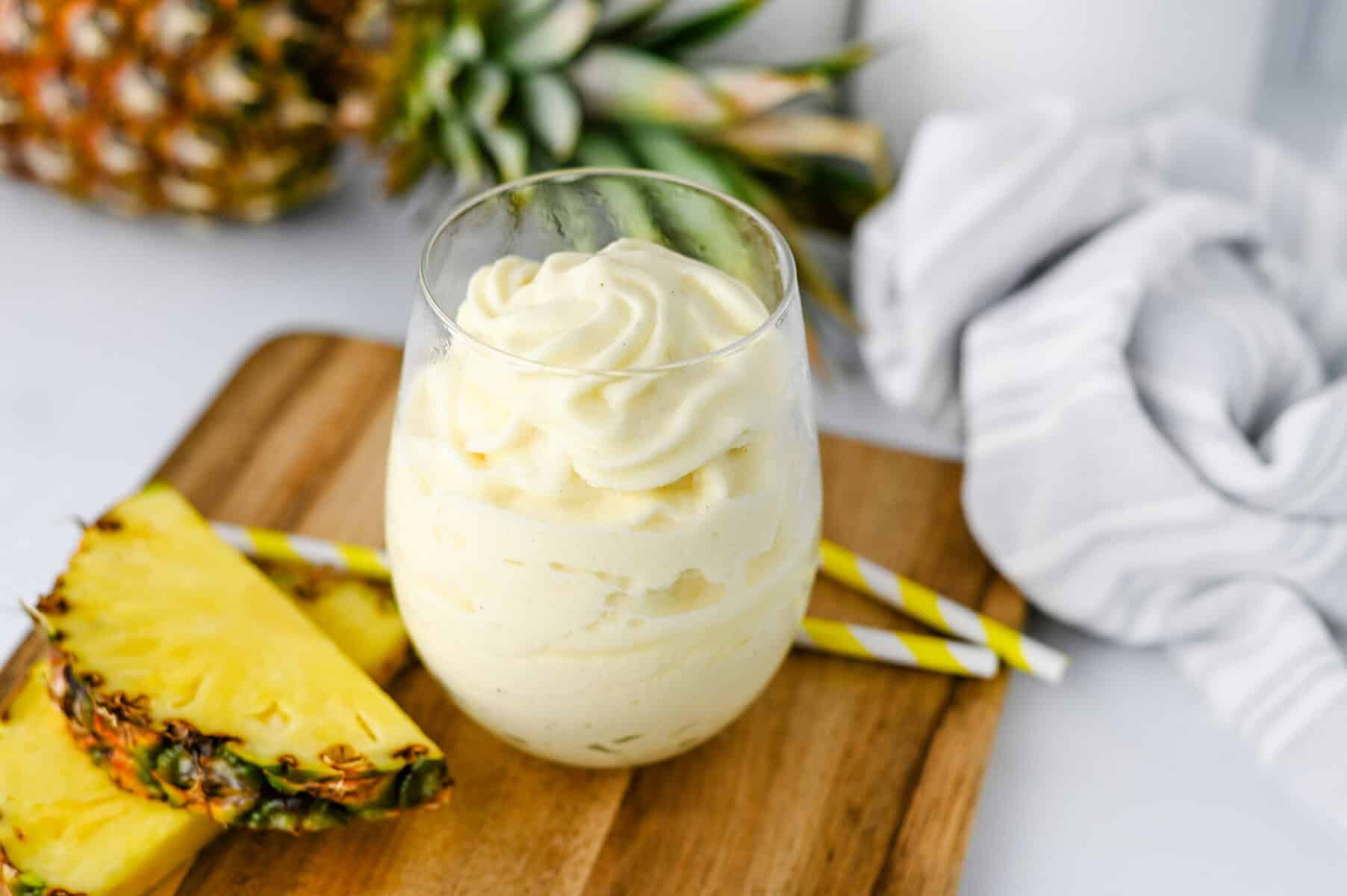 A glass of creamy pineapple dessert sits on a wooden board, garnished with two yellow-striped straws and pineapple slices, evoking the classic Disney Dole Whip Recipe. A whole pineapple and a white cloth are in the background.