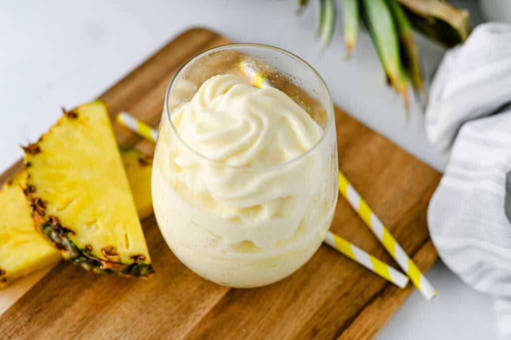 A glass of creamy pineapple soft serve, inspired by the Disney Dole Whip Recipe, sits on a wooden board next to two pineapple slices and yellow-striped straws. A green pineapple crown is partially visible in the background.