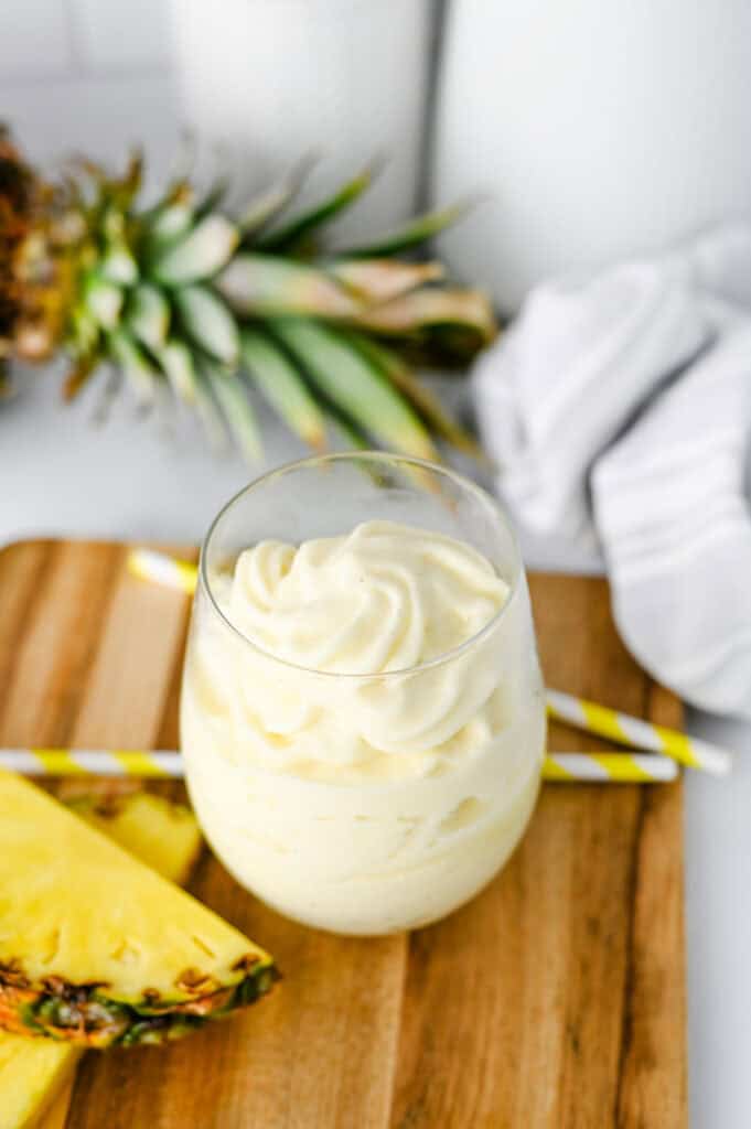 A glass filled with creamy, swirled pineapple dessert sits on a wooden board, inspired by the classic Disney Dole Whip Recipe, with a fresh pineapple, a wedge of pineapple, and two yellow-striped straws nearby.