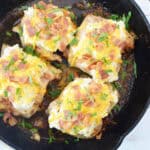 Four Cheesy Ranch Chicken breasts baked in a black cast iron skillet, topped with melted cheese, chopped bacon, and fresh herbs, sitting on a white wooden surface.