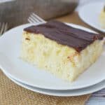 A slice of chocolate eclair poke cake on a white plate with forks nearby and another slice in the background.