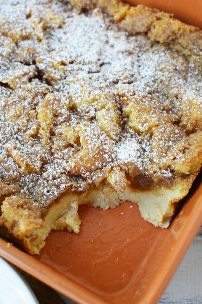 A baked Overnight French Toast Casserole in an orange-brown dish, topped with powdered sugar. A portion has been scooped out, revealing the soft, moist texture inside.